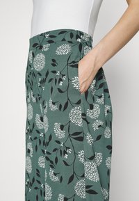 Saia verde com padrão floral, com cintura elástica e bolsos laterais. Apresenta desenhos de flores em branco e preto e uma textura suave.