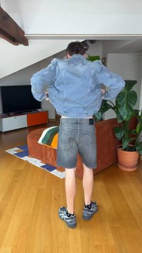 Άτομο που φοράει τζιν μπουφάν και σορτς στέκεται με την πλάτη στην κάμερα σε σαλόνι με πορτοκαλί καναπέ και μεγάλο γλάστρα φυτό.