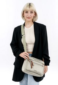 Beige křížová taška vyrobená z odolné látky, s více zipovými přihrádkami, s hnědými koženými detaily a měkkým povrchem.