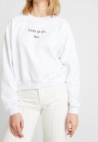 Sweatshirt blanc à col rond, avec des manches raglan et des poignets côtelés. Avec le texte noir "D'abord, non." centré sur la poitrine.