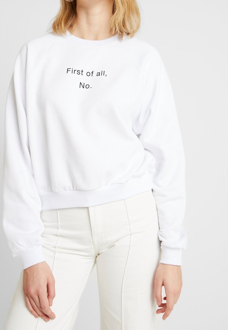 Sweatshirt blanc à col rond, avec des manches raglan et des poignets côtelés. Avec le texte noir "D'abord, non." centré sur la poitrine.