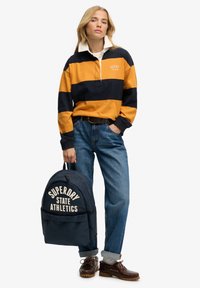 Langærmet poloshirt med striber i orange og marineblå, med en hvid krave. Blå jeans, brune sko og en marineblå rygsæk med hvid tekst, der lyder "SUPERDRY STATE ATHLETICS."