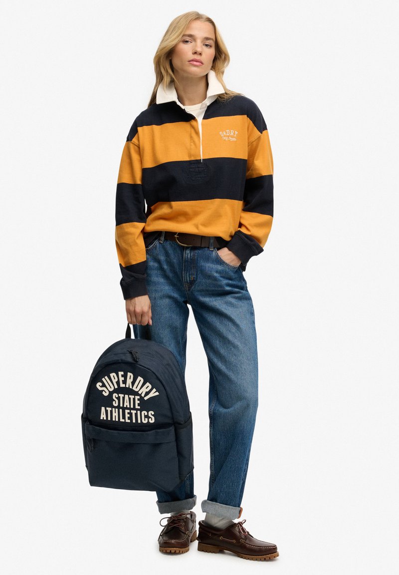 Langærmet poloshirt med striber i orange og marineblå, med en hvid krave. Blå jeans, brune sko og en marineblå rygsæk med hvid tekst, der lyder "SUPERDRY STATE ATHLETICS."