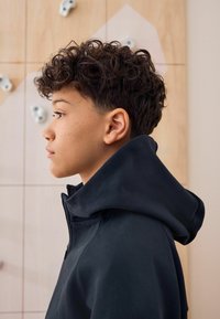 Zwarte rits-hoodie met een hoge kraag, gladde stof en een grote capuchon. Krullend haar en sproeten zichtbaar op het model tegen een klimwand.