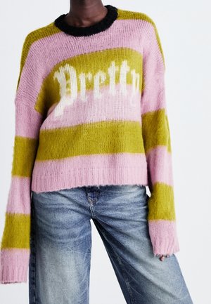 Personne portant un pull duveteux rayé rose et vert avec le texte « Pretty » sur le devant et un jean bleu délavé sur un fond blanc uni.