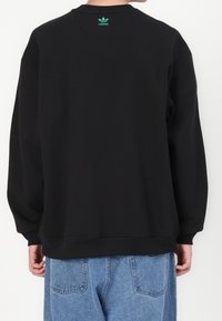 Svart sweatshirt med avslappnad passform, ribbade ärmslut och nederkant, samt liten grön Adidas-logga på baksidan av nacken. Buren med ljusblå jeans.