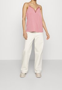 Camisola rosa con rifiniture in pizzo e scollatura a V, abbinata a pantaloni bianchi larghi e scarpe beige. Texture piatta e liscia.