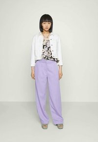 Uma jovem mulher está de pé, vestindo um casaco branco abotoado, uma blusa preta com estampado floral, calças largas lilás e sandálias de plataforma verdes, contra um fundo neutro.