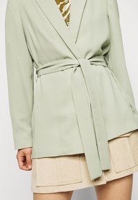 Ljusgrön kavaj med knytband i midjan, med slät tygkvalitet och långa ärmar, tillsammans med en beige strukturerad kjol.