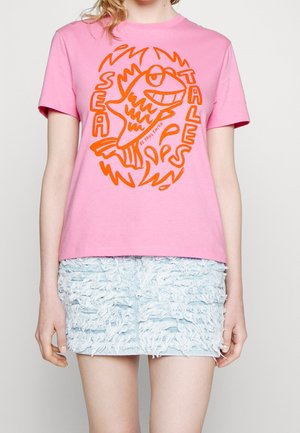 Person iført en pink T-shirt med en orange stiliseret fisk og ordene "Sea Tales" kombineret med et lyseblåt tekstureret mini-nederdel.