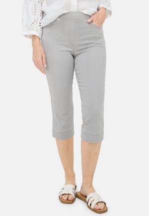 Femme portant un pantalon court gris clair et des sandales blanches à enfiler avec un chemisier blanc, une main dans la poche, sur un fond uni.
