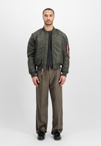 Olive green bomber jacket with ribbed cuffs, zip closure, and patch detail on sleeve, paired with beige pleated trousers and black shoes.