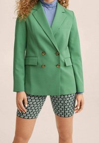 Zielona podwójnie zapinana marynarka z złotymi guzikami, noszona na jasnoniebieskim golfie. Połączona z czarnymi szortami rowerowymi w kwiatowy wzór.