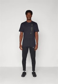 Melnas Nike T-krekls ar logotipu un grafiskām detaļām priekšpusē, kombinēts ar melnām koniskām biksēm un melnām sporta kurpēm.