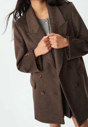 Vrouw draagt een dubbelgeknoopte bruine wollen jas over een grijze gebuttonde trui en houdt de jas met beide handen dicht.