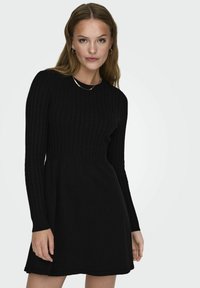 Robe noire côtelée à manches longues, corsage ajusté et jupe évasée. Fabriquée en matériau doux avec un décolleté rond et une texture subtile.