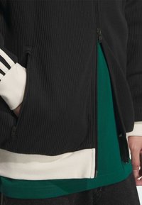 Crna rebrasta jakna sa patentnim zatvaračem, bijelim rukavima i zelenom majicom ispod; ima patentni zatvarač, džepove i kontrastni blok boje na dnu.