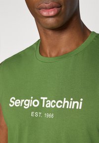 Grøn bomulds ærmeløs t-shirt med hvid tekst "Sergio Tacchini EST. 1966" centreret på fronten, der fremhæver en glat tekstur.