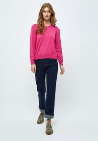 Maglione rosa con maniche lunghe e orlo a coste, abbinato a jeans blu scuro arrotolati alle caviglie, e mocassini marroni con dettagli sottili.