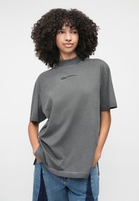 Šedé tričko s krátkym rukávom a vysokým golierom, s logom v čiernej farbe. Vyrobené z mäkkej tkaniny s voľným strihom, s hladkou textúrou.