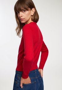 Maglione rosso lavorato a maglia con scollatura rotonda e maniche lunghe, con polsini e orlo a costine. Abbinato a jeans in denim a vita alta.