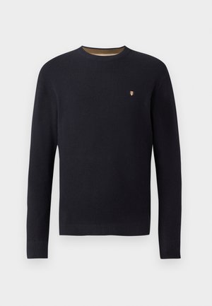 Marineblå strikkegenser med rund hals, ribbestrikkede mansjetter, og et lite emblem på brystet. Teksturert stoff med et subtilt mønster.