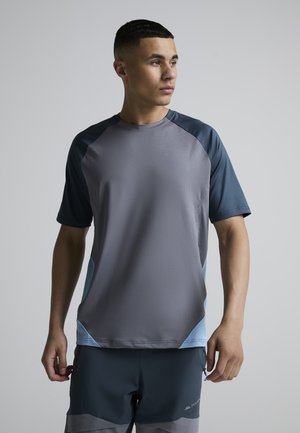 Ung mand i grå og blå atletisk t-shirt med korte ærmer og mørke shorts, der kigger til venstre mod en ensfarvet baggrund.