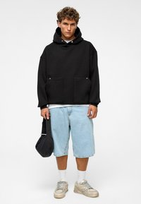 Zwarte oversized hoodie met een trekkoord, voorzien van twee voortassen, gecombineerd met lichtblauwe denimshorts, witte sokken en sneakers.