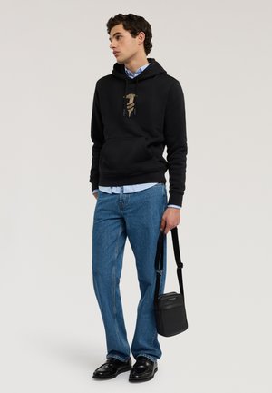Młody mężczyzna w czarnej bluzie z kapturem z nadrukiem, niebieskich dżinsach i czarnych butach, trzymający czarną torbę na ramię za pasek.