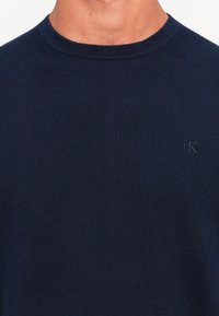 Pulover bleumarin cu guler rotund, realizat dintr-o țesătură cu model în relief, având un logo brodat subtil pe piept. Croială clasică, fără modele sau accente.
