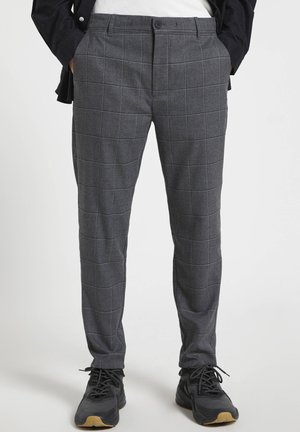 Man draagt grijze geruite slim-fit broek en zwarte sneakers met lichtbruine zolen, handen in de zakken, staand op een witte achtergrond.