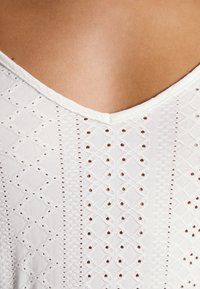 Gros plan d’un vêtement en tissu œillet blanc avec des motifs en losange et points porté sur la peau avec un col en V.