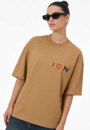 Femme portant des lunettes de soleil noires et un t-shirt marron oversize avec "I heart you" brodé sur le devant, associé à un jean bleu.