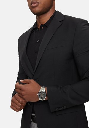 Juodas bleizeris su atvartais, derinamas su juodu polo marškinėliais. Ant rankos sidabrinis laikrodis su tamsiu ciferblatu ir trimis subdials. Slygi tekstūra.