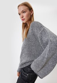 Grau gestrickter Pullover mit lockerer Passform, dicker Rippstruktur und weiten Ärmeln, kombiniert mit dunklen Jeans. Dezenter Kontrast in den Farbtönen.