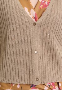 Beige Rippstrick-Cardigan mit V-Ausschnitt, ausgestattet mit Knopfleiste und dezenten Glitzerakzenten. Über einer blumenmusterierten Bluse in warmen Farbtönen getragen.