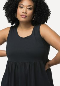 Donna con capelli ricci che indossa orecchini a cerchio e un vestito nero senza maniche con vita arricciata, in piedi con le mani sui fianchi.