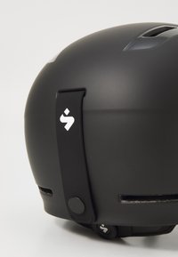 Matna črna čelada z ventilacijskimi režami, nastavljivim plastičnim trakom in detajlom logotipa; ima okroglo obliko in gladko teksturo.