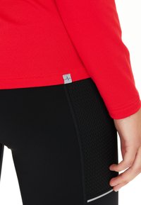 Rotes langärmeliges Oberteil kombiniert mit schwarzen Leggings, die ein strukturiertes Paneel, reflektierende Akzente und ein Logo-Tag in der Nähe des Saums aufweisen.
