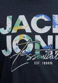 Crna majica s bold bijelim natpisom i višebojnim mrljama boje. Natpisi uključuju "JACK & JONES", "Essential" i "EST. 1990."