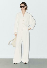 Veste blanche sur mesure avec des boutons dorés et un pantalon large blanc. Les accessoires incluent des lunettes de soleil oversize noires et un sac beige. Texture lisse.