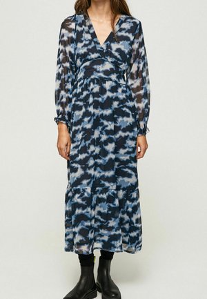 Kobieta ubrana w długą sukienkę z długim rękawem i dekoltem w serek, o długości do kostek, z niebiesko-czarnym abstrakcyjnym wzorem chmury oraz czarne botki za kostkę.