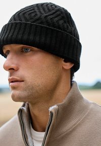 Melna adīta cepure ar tekstuētu rakstu, ribota aproce un cieša piegulšana. Valkāta kopā ar bēšas krāsas zip-up džempers, ar trikotāžas oderi.