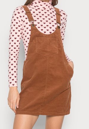 Vrouw draagt een bruine corduroy tuinbroekjurk met voorzak over een roze longsleeve met patroon van kleine rode hartjes.