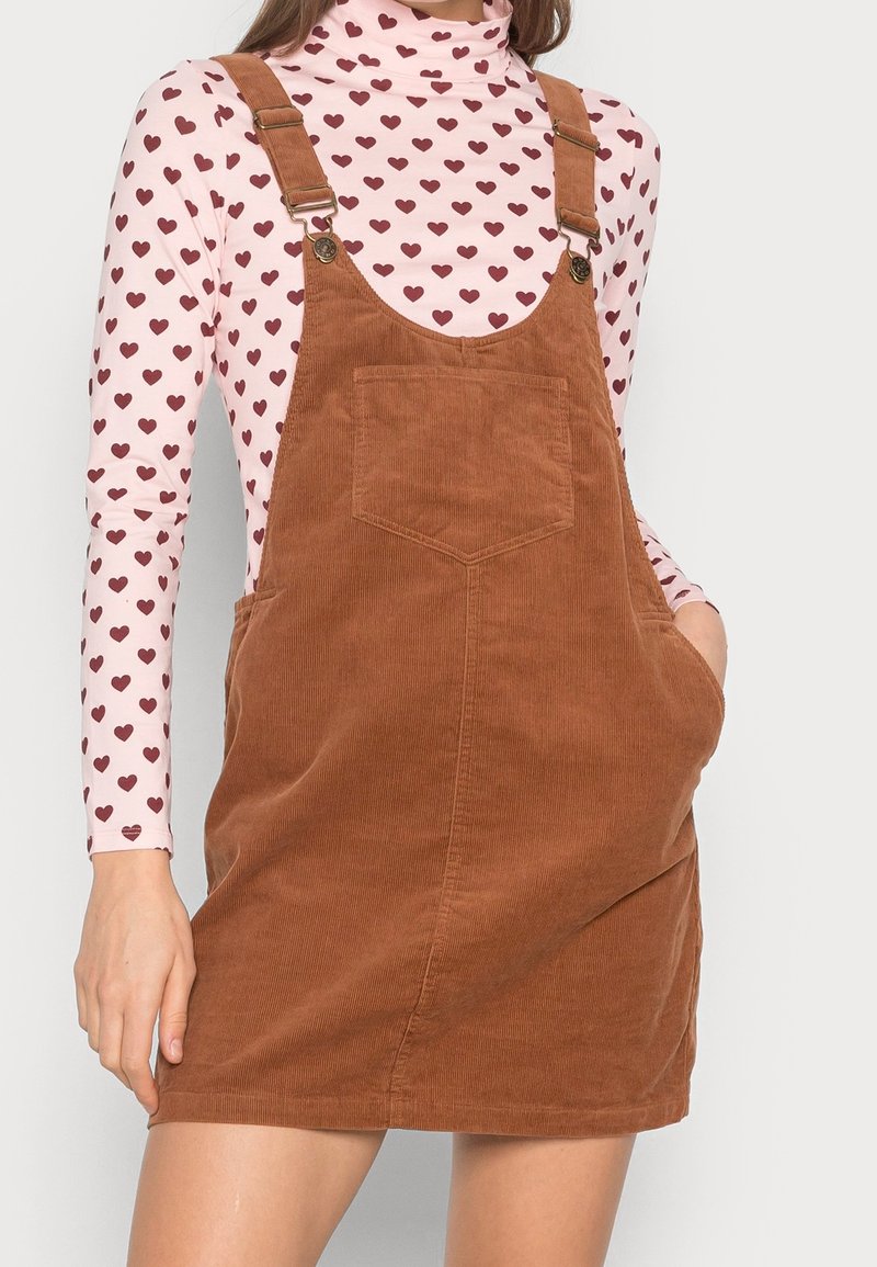 Femme portant une robe salopette en velours côtelé marron avec poche avant sur un haut rose à manches longues à motif de petits cœurs rouges.