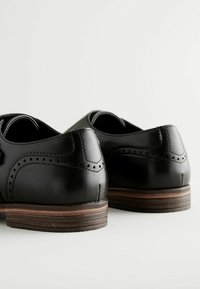 Crne kožne cipele s glatkom završnom obradom, brogue detaljima i složenim drvenim potplatom. Dizajn uključuje zaobljeni stražnji dio i bočno šivanje.