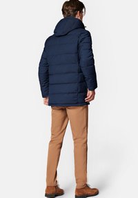 Granatowa kurtka puchowa z kapturem, z pikowanym wzorem i zamek błyskawiczny. Beżowe spodnie i brązowe buty uzupełniają stylizację.