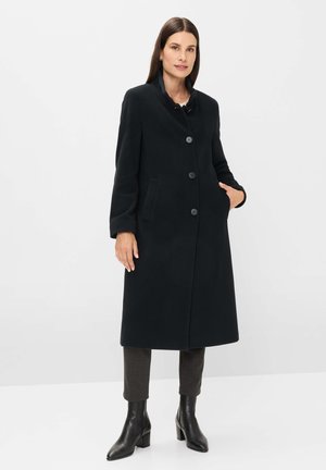 Frau mit glattem braunem Haar, die einen langen schwarzen Mantel mit Knöpfen, dunkle Hosen und schwarze Lederstiefeletten trägt und vor einem weißen Hintergrund steht.