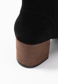 Botte à talon haut en daim noir avec un talon en bloc brun texturé et une semelle noire lisse. Présente des coutures visibles le long de la couture.