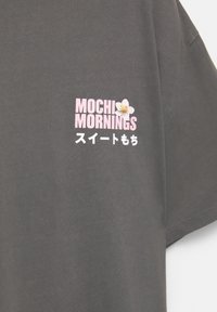 T-shirt en coton gris avec le texte rose "MOCHI MORNINGS" et un petit graphique de fleur, accompagné de caractères japonais en dessous. Design à manches courtes.